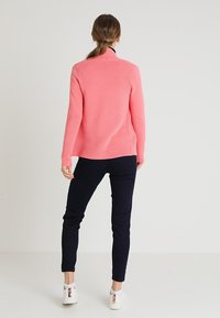 Różowy sweter z prążkowanej wełny z wysokim kołnierzem, połączony z ciemnogranatowymi spodniami typu slim fit i białymi trampkami. Tekstura wydaje się miękka i przytulna.