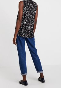 Schwarze, ärmellose Bluse mit floralem Muster und hohem Kragen, kombiniert mit blauen Jeanshosen mit umgeschlagenen Säumen und schwarzen Slippern.