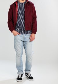 Chaqueta burdeos con forro de cuadros rojos sobre una camisa de patrón azul marino. Jeans de mezclilla azul claro y zapatillas negras con detalles blancos. Atuendo casual.