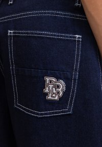 Un pantalon en denim bleu foncé présente une poche arrière avec des coutures blanches contrastantes et un logo brodé en beige et blanc.