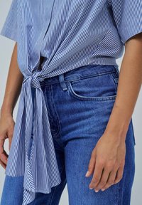 Camicetta a righe blu e bianche legata in vita con un nodo, abbinata a jeans in denim blu scuro. La camicetta presenta piccoli dettagli decorativi.