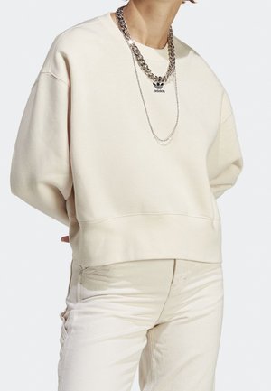 Osoba ubrana w kremową bluzę Adidas z czarnym logo, dopasowane jasne spodnie, dopełnione dwoma srebrnymi łańcuszkami.