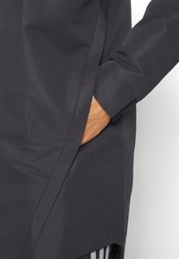 Main d'une personne dans la poche d'un manteau noir à manches longues, montrant la couture latérale du manteau et la texture lisse du tissu.