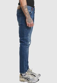 Jean skinny en denim bleu avec des détails usés, taille mi-haute, et un patch en cuir à l'arrière de la taille. Associés à des chaussures de sport argentées.
