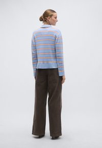 Μπλε ριγέ πουλόβερ με πορτοκαλί και κίτρινες ρίγες, μακριά μανίκια και γιακά, σε συνδυασμό με καφέ παντελόνι φαρδύ.