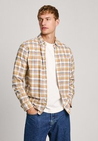 Camicia a quadri marrone e beige con dettagli bianchi, indossata sopra una maglietta bianca. Modello che indossa jeans di denim blu, mani in tasca.