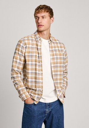 Camicia a quadri marrone e beige con dettagli bianchi, indossata sopra una maglietta bianca. Modello che indossa jeans di denim blu, mani in tasca.