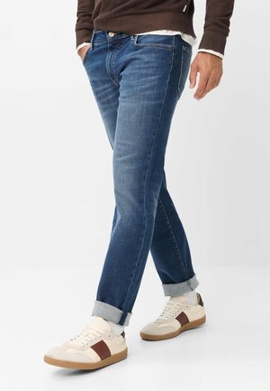 Uomo con jeans blu risvoltati, calzini bianchi, sneakers beige con strisce marroni e maglione marrone con le mani in tasca.