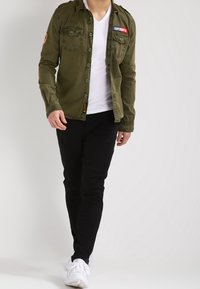 Giacca bottonata verde oliva con due tasche sul petto e toppe, indossata su una maglietta bianca a scollo a V, abbinata a pantaloni neri e sneakers bianche.