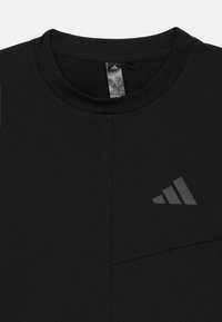 Melnā sporta T-krekls ar apaļu izgriezumu, izgatavots no gludas auduma. Kreisajā krūtīs ir reljefs, tonēts logotips un redzama šuve.