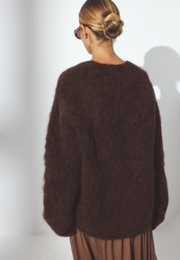 Brown fuzzy sweater with a loose fit and dropped shoulders, featuring a soft texture and solid color. Worn over a pleated brown skirt.
