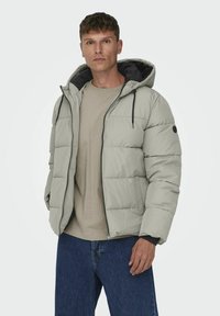 Ljusgrå pufferjacka med huva, dragkedja framtill och svarta accenter. Tjock, vadderad design med innerfoder och sidofickor.