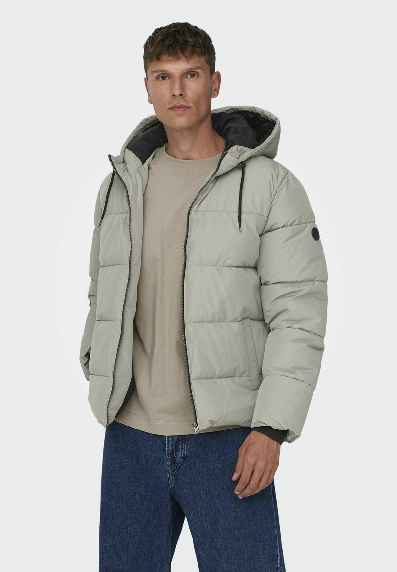 Ljusgrå pufferjacka med huva, dragkedja framtill och svarta accenter. Tjock, vadderad design med innerfoder och sidofickor.