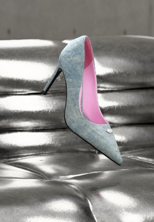 Spids tå højhælet sko med denim-lignende tekstur, med pink inderside og sølvfarvet detalje, hvilende på en skinnende sølvoverflade.
