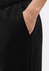 Χέρι ακουμπισμένο στην εμπρόσθια τσέπη μαύρου αθλητικού παντελονιού Ted Baker με ελαστική μέση, κοντινή λήψη υφάσματος και λογότυπου.