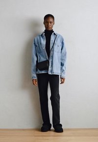 Veste en denim bleu clair, col roulé noir, pantalon noir et chaussures foncées. Sac bandoulière en bandoulière. Sol en bois, fond uni.