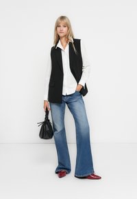 Un gilet sans manches noir sur une chemise blanche à boutons, associé à un jean bleu évasé et des chaussures rouges à bout pointu. Les accessoires incluent un petit sac noir.