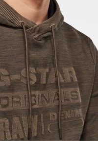 Hnědá mikina s kapucí s žebrovanou texturou, stahovacím šňůrkem a vyraženými logy. Tkanina působí měkce a lehce opotřebovaně.