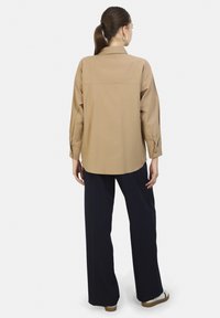 Beige langærmet shirt met een rugpand en geknopte manchetten, gecombineerd met wijde marineblauwe broek en lichte instappers. Gladde stoftextuur.