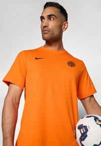 Camiseta Nike naranja con cuello redondo y mangas cortas, que presenta un logo negro en el pecho. Se sostiene un balón de fútbol con un diseño en el brazo.