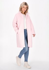 Cappotto in pile rosa chiaro con zip frontale, tasche laterali e cappuccio. Il modello indossa jeans aderenti e sneakers beige. Materiale morbido e strutturato.