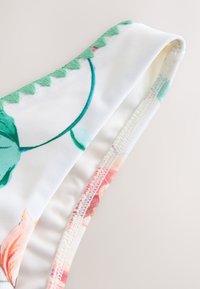 Cvetlične bikini hlačke z belo osnovo, okrašene z zelenimi listi in rožnatimi cvetličnimi vzorci. Robovi so obrobljeni z zelenimi volančki in gladko teksturo.