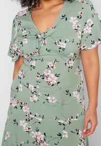 Frau in einem grünen Kleid mit pink-weißen Blumenmustern und einem Frontknoten, das einen Teil ihres Oberkörpers und ihrer Arme zeigt.