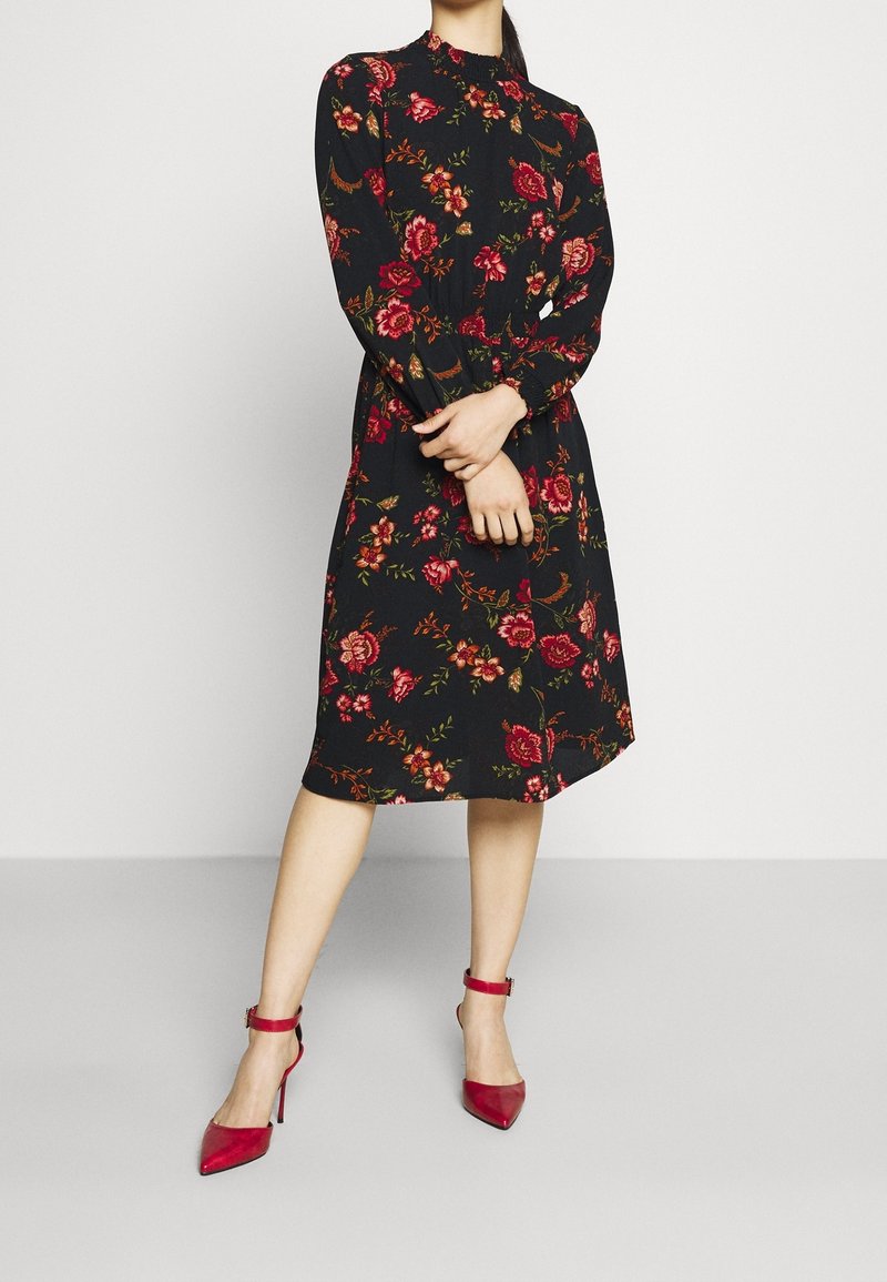 Robe florale noire à manches longues, avec un col à volants et des motifs de roses rouges. Associée à des chaussures à talons rouges avec des lanières à la cheville.