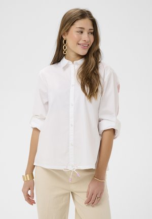 Jeune femme regardant sur le côté, portant une chemise blanche boutonnée, un pantalon beige et des bijoux en or, avec de longs cheveux ondulés.