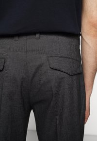 Närbild av en person som bär mörkgrå, texturerade byxor med fickor bak och en svart skjorta instoppad.