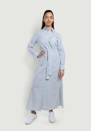 Donna in piedi con la mano sul fianco, indossa un lungo vestito a righe azzurre con bottoni, cintura e sneakers bianche su uno sfondo semplice.