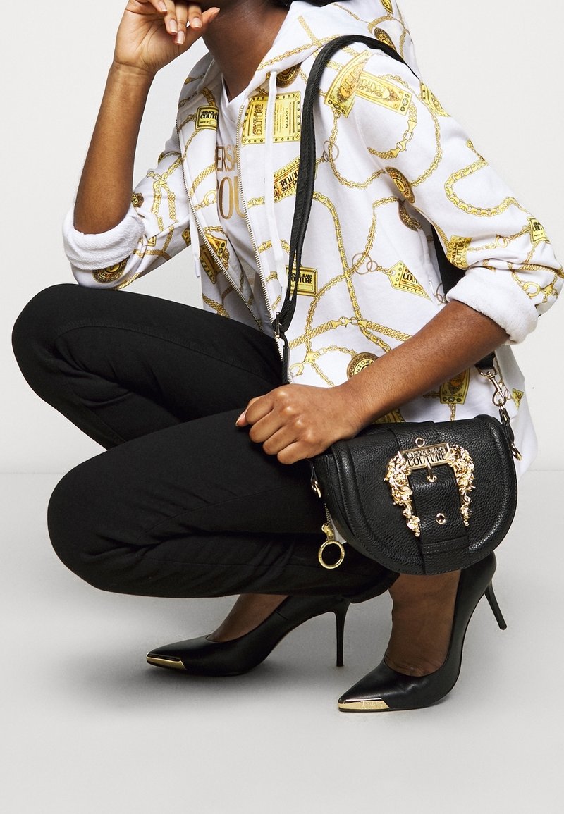 Bolso negro texturizado con herrajes dorados y acentos de cabeza de león, combinado con pantalones negros ajustados y tacones negros puntiagudos con puntas doradas.
