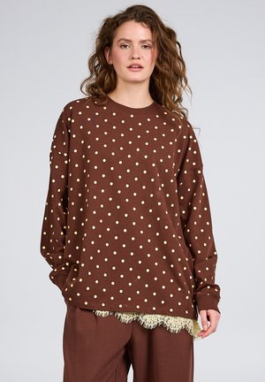 Kvinde med krøllet hår iført en brun langærmet skjorte med beige polkaprikker og matchende bukser med blonder i taljen.