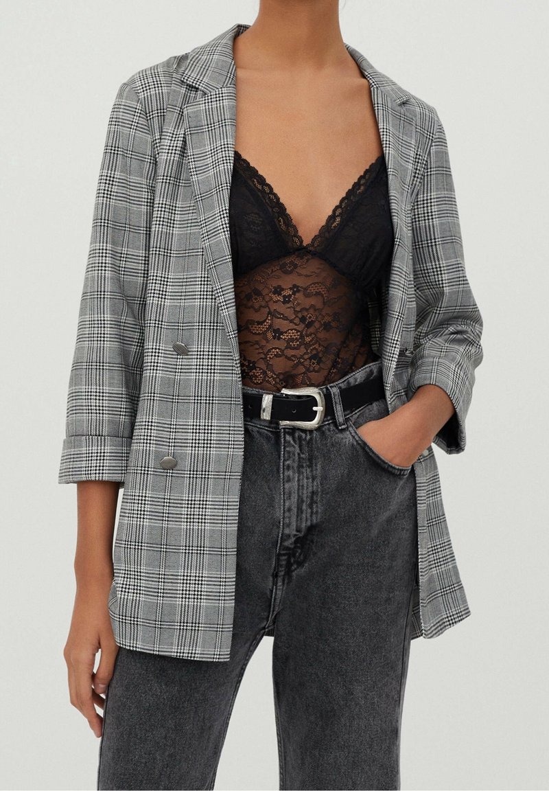 Femme portant un blazer gris à carreaux, un caraco en dentelle noire, un jean noir taille haute et une ceinture noire avec une boucle argentée.