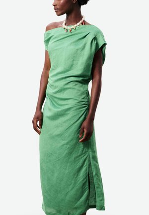 Femme portant une robe verte à épaules dénudées avec une fente latérale et un collier de perles avec des perles colorées, debout devant un fond uni.