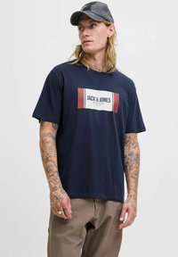 Sötétkék, rövid ujjú póló, elülső részén egy téglalap alakú, fehér és piros grafikai logóval, amelyen a 'JACK & JONES' felirat szerepel, bézs nadrággal párosítva.