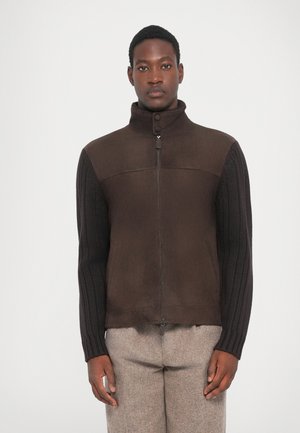 Braune Wildlederjacke mit gerippten Strickärmeln; verfügt über einen hohen Kragen und einen Reißverschluss, der einen strukturierten Materialkontrast zeigt.