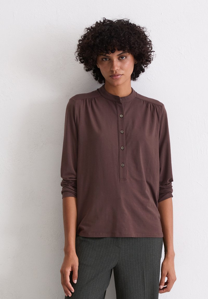 Chemise marron à manches longues avec un col haut, détails de boutons et texture douce, assortie à un pantalon gris à rayures.