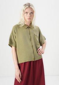 Olijfgroen shirt met korte mouwen, een rits aan de voorkant en zak, met een relaxte pasvorm en strakke lijnen, gecombineerd met een bordeauxrode rok.