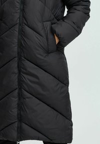 Abrigo acolchado negro con diseño de rombos, cierre de cremallera y bolsillos laterales. Presenta un exterior suave y una textura acolchada.