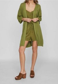 Cardigan en maille vert olive porté sur une combinaison assortie verte ; présente un décolleté en V et des manches amples. Sandales à plateforme en bois marron.