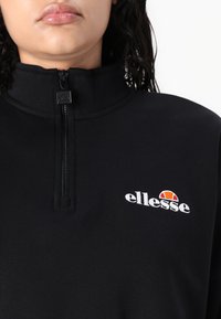Felpa nera con collo alto e chiusura con zip. Presenta un logo ricamato e un tessuto liscio e strutturato. Design semplice e minimalista.