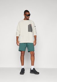 Sweatshirt in off-white with a textured dark pocket, paired with teal shorts. Black sneakers and sunglasses complete the outfit.