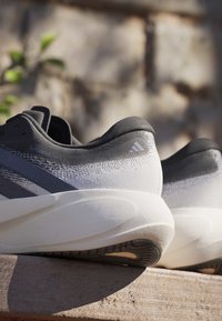 Paire de chaussures de course Adidas Supernova Rise 3 noires et blanches posées sur une surface en bois avec un arrière-plan extérieur flou.
