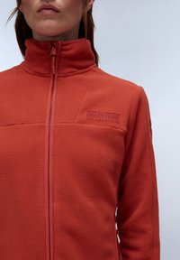 Giacca in fleece arancione con colletto alto, zip completa e piccolo logo a patch sul petto. Presenta una texture liscia e un design aderente.