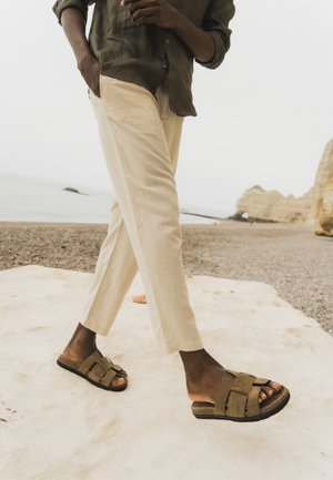 Persona che indossa pantaloni beige e sandali marroni che cammina su una superficie di pietra chiara con una spiaggia rocciosa e il mare sullo sfondo.