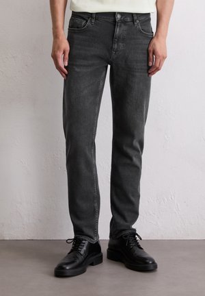 Persoon in slim-fit vervaagde zwarte jeans en zwarte leren veterschoenen staat op een grijze vloer tegen een witte muur.