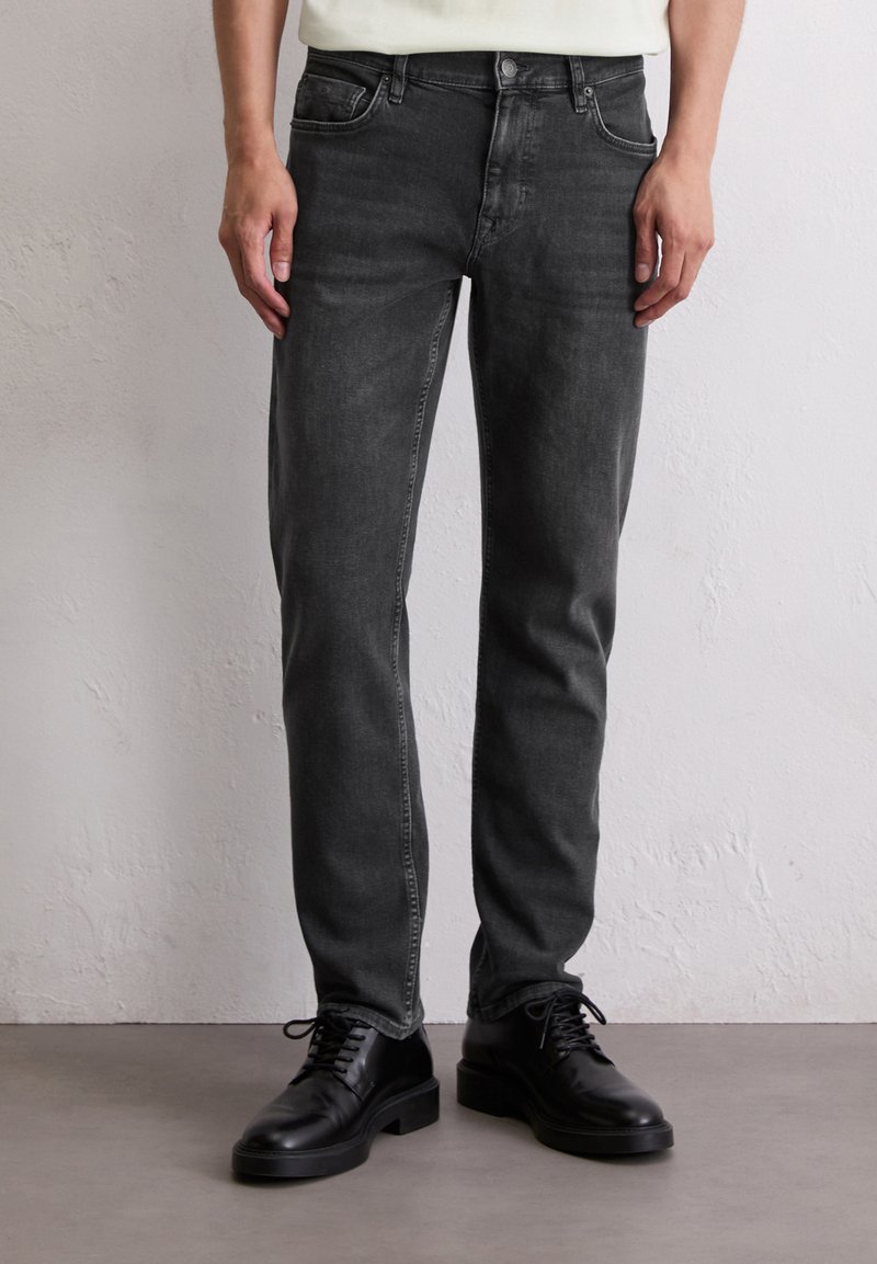 Persoon in slim-fit vervaagde zwarte jeans en zwarte leren veterschoenen staat op een grijze vloer tegen een witte muur.