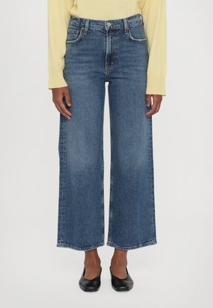 Højttaljede vidde-ben jeans i mørkeblå denim med rå kant, som har fem lommer og en knaplukning. Modellen har sorte ballerinaer på.