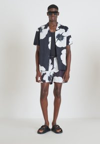 Camicia a maniche corte con motivo floreale in bianco e nero, abbinata a pantaloni corti coordinati. Modello che indossa occhiali da sole neri e sandali.